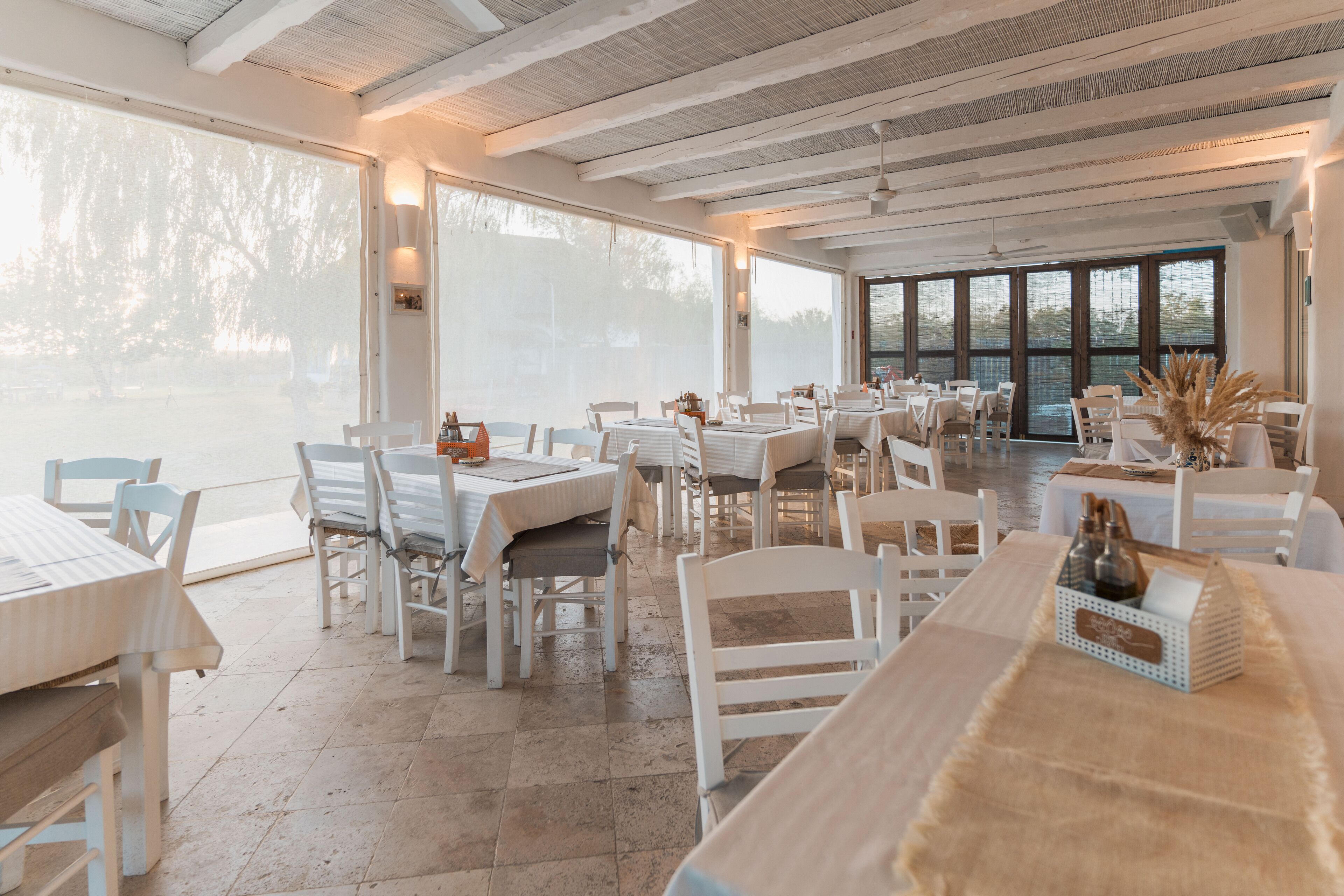 A spacious restaurant with white tables and chairs, large windows, and a view of the outdoors.