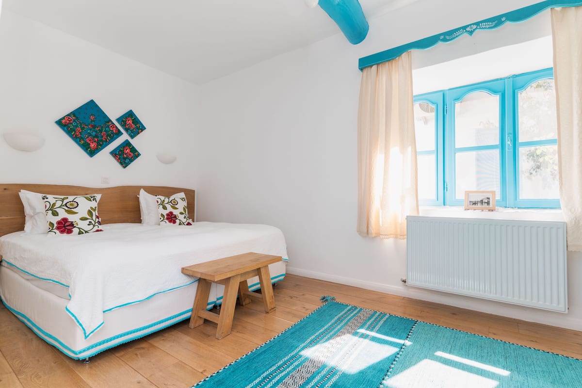A bedroom with a bed, a wooden bench, a blue and white patterned rug, and a window with curtains.
