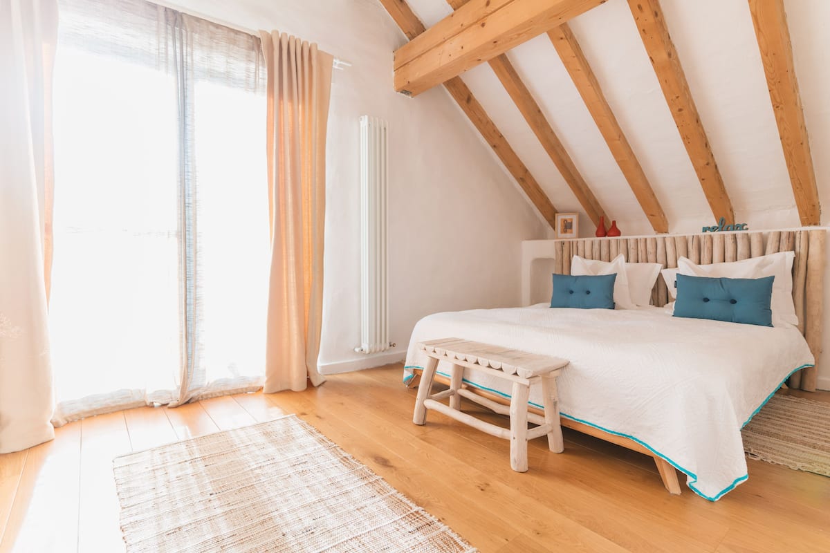 A bedroom with a wooden ceiling, a bed with white bedding and blue pillows, a small wooden stool, and a window with curtains.