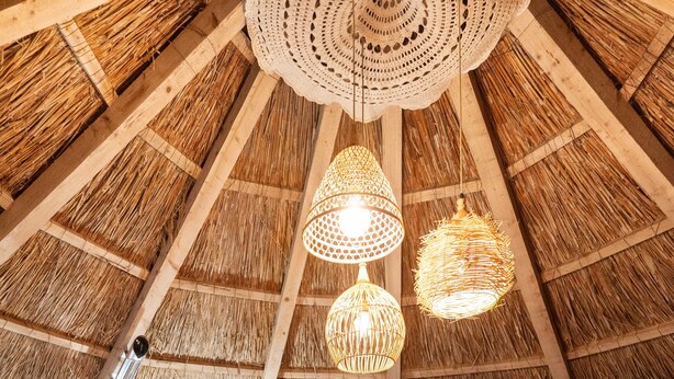 Un interior con techo de paja, mobiliario de mimbre, una lámpara de pie trenzada y un arreglo floral central.