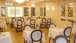 Uma sala de jantar formal com várias mesas e cadeiras, lustres e grandes janelas.