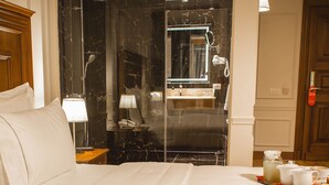 Quarto de hotel com uma cama grande, uma mesa de cabeceira com abajur, banheiro com espelho e um serviço de chá e pastelaria.