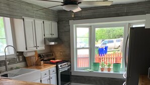 Una cocina con una isla central, armarios blancos, una estufa y un ventanal con vistas a una valla roja y vehículos afuera.