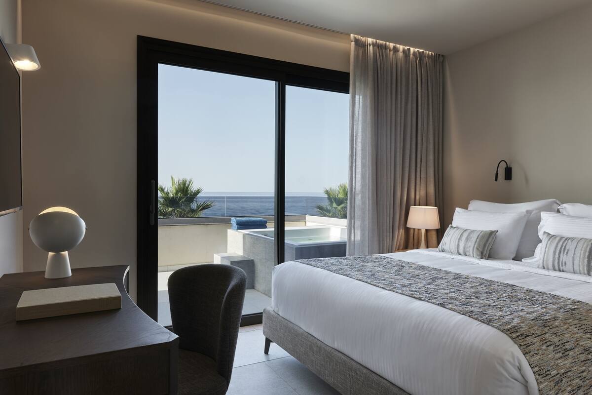 A modern hotel room with a large bed, a desk with a lamp, a chair, and a view of the ocean through a sliding door.