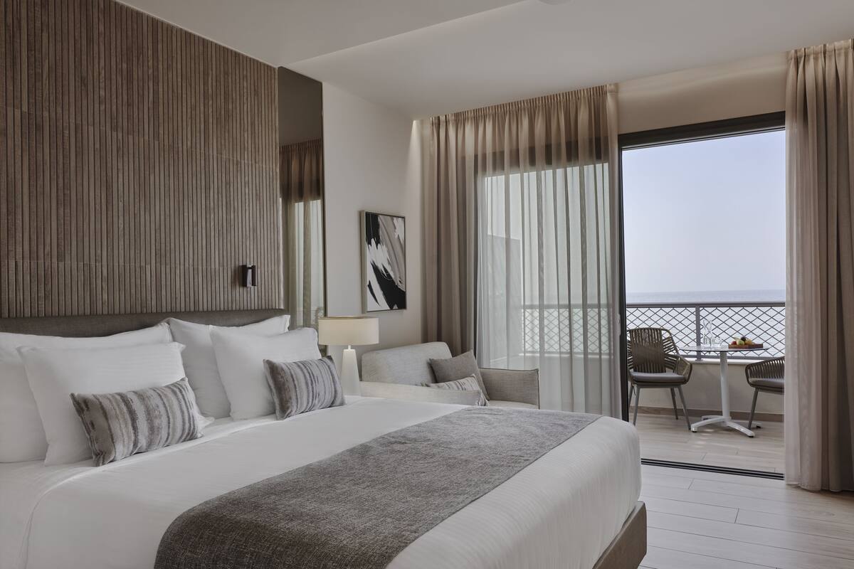 A modern hotel room with a large bed, a balcony with chairs, and a view of the sea.