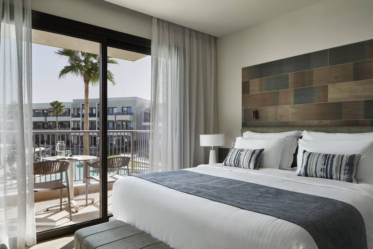 A hotel room with a large bed, a balcony with a pool, and a view of palm trees and buildings.