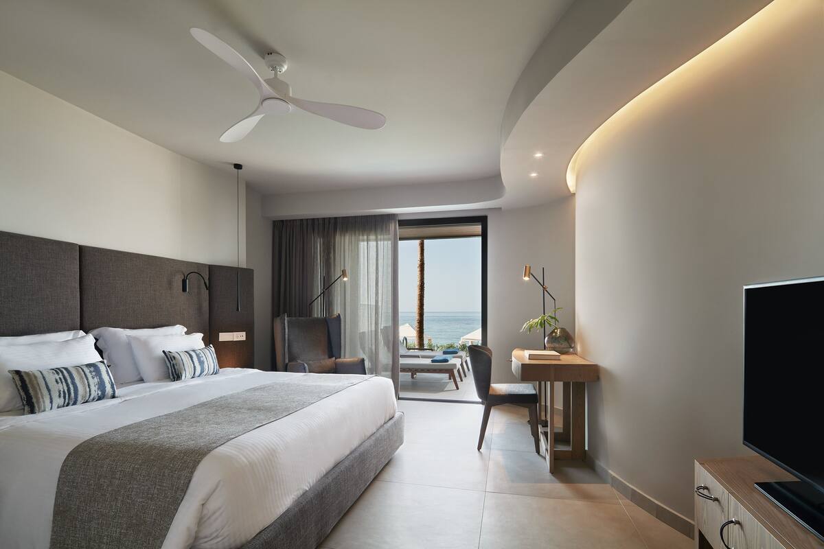 A modern hotel room with a large bed, a desk, a TV, and a balcony with a view of the ocean.
