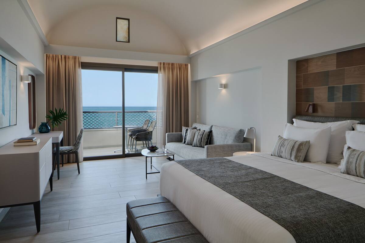 A modern hotel room with a large bed, a desk, a chair, and a balcony with a view of the sea.