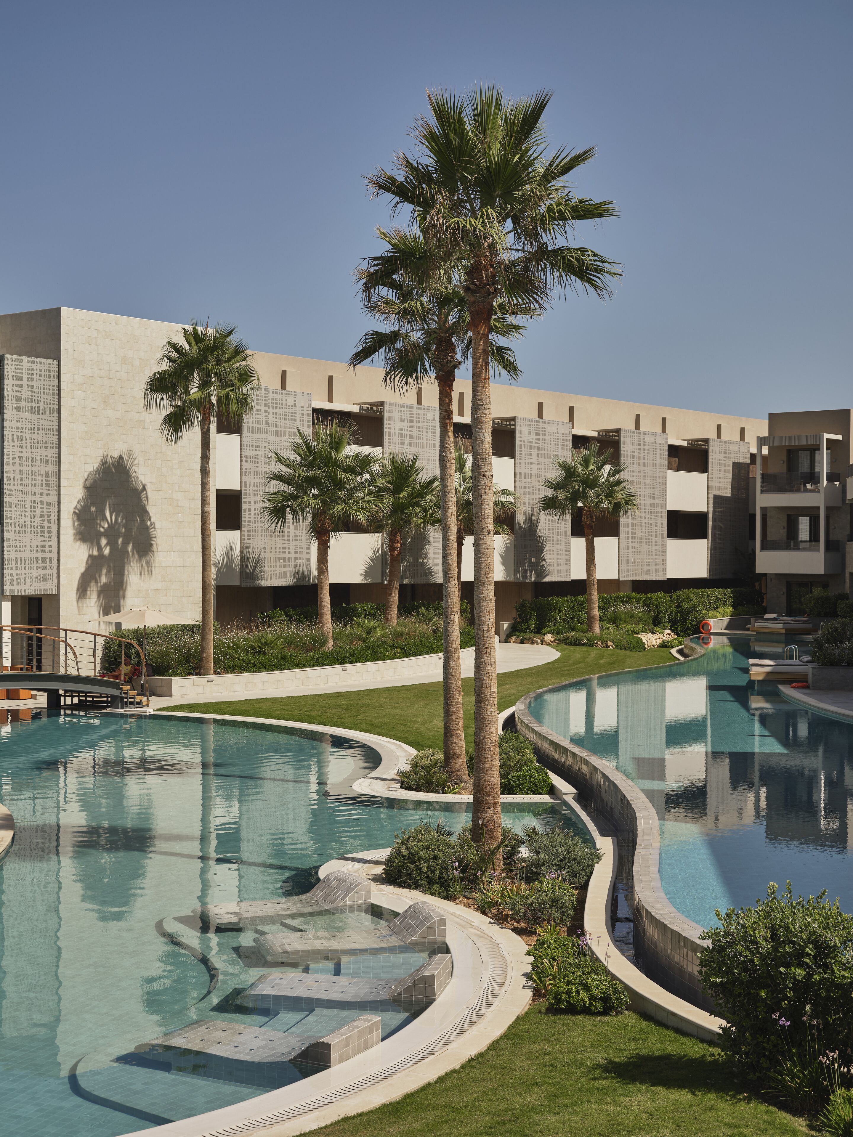 Een modern hotel met een groot zwembad, palmbomen en een helderblauwe lucht.