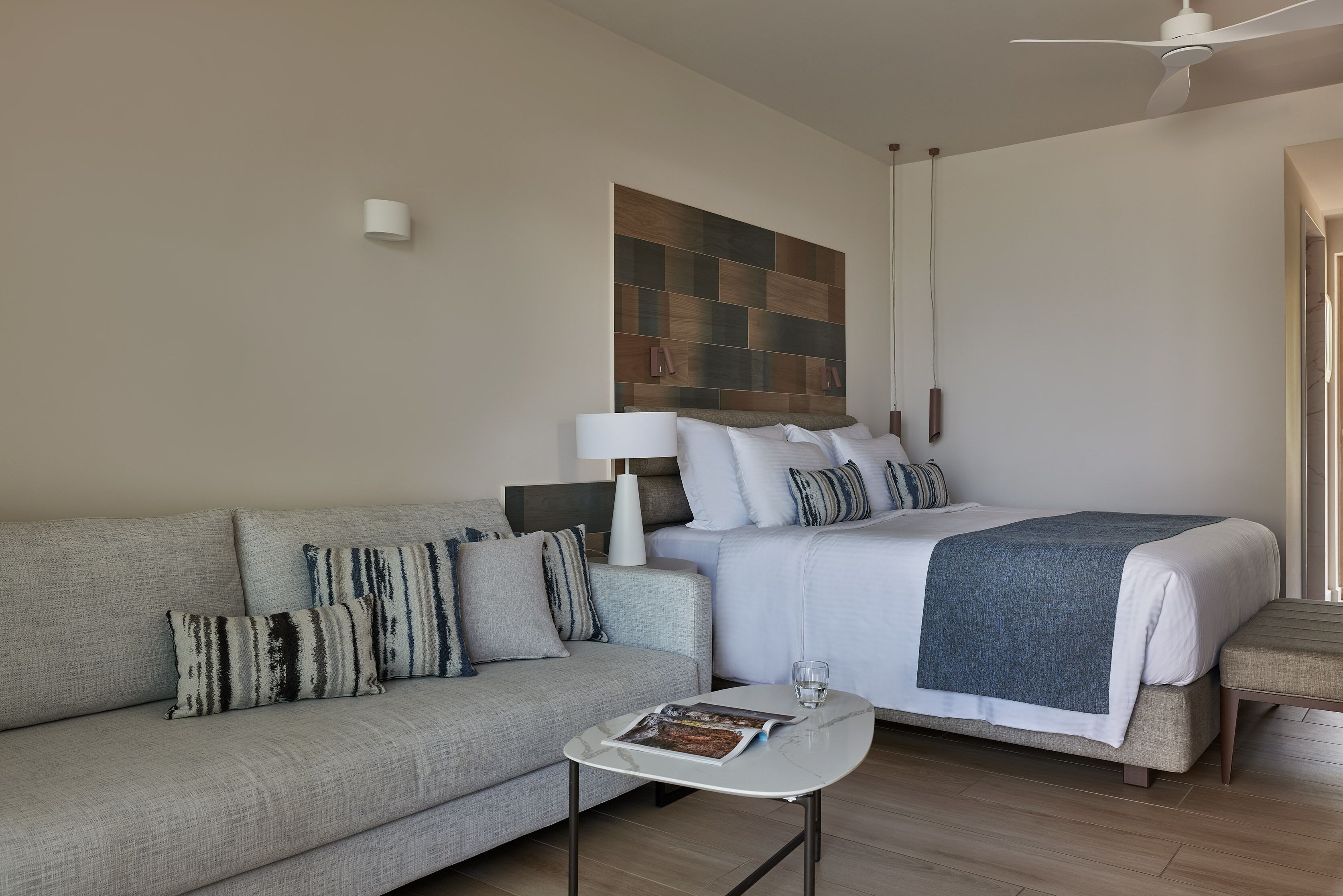 Una habitación de hotel moderna con un sofá, una cama con una manta azul, una mesita con una revista y un ventilador de techo.
