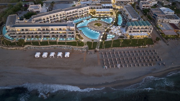 An aerial view of a resort with a large pool, multiple buildings, and a beach area with umbrellas and lounge chairs. - AMIRA LUXURY RESORT & SPA (Rethymno)