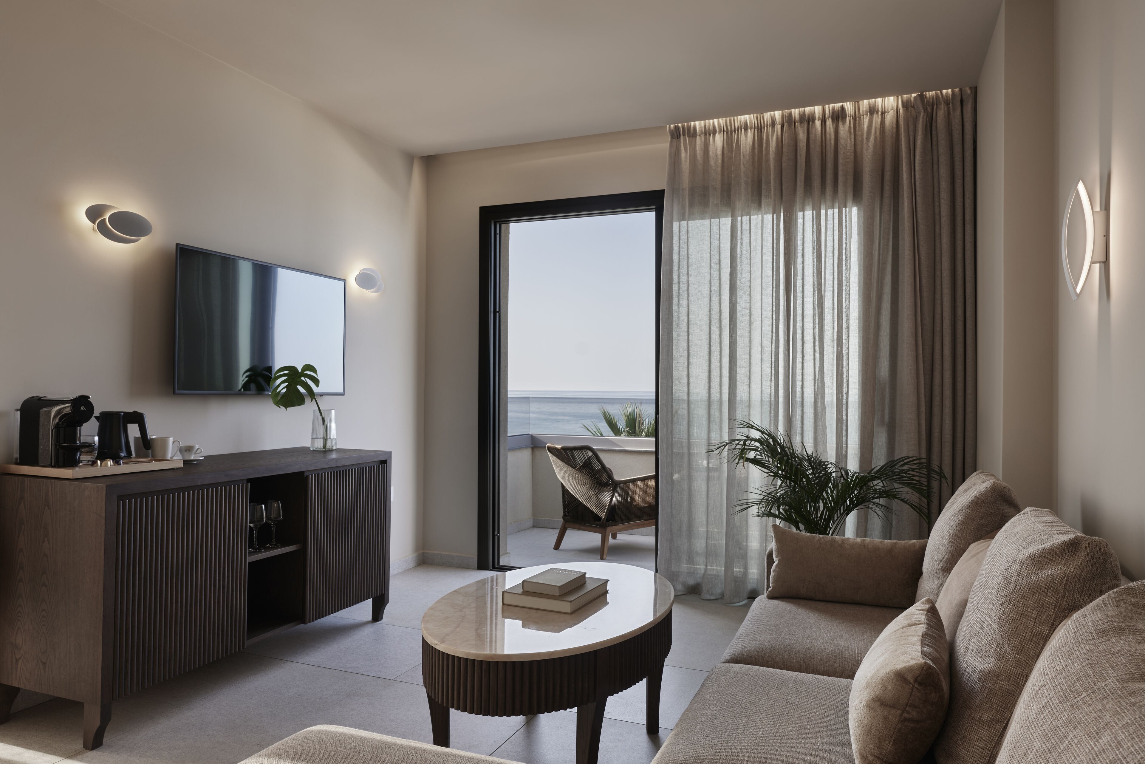 A modern living room with a sofa, a coffee table, a TV, and a balcony with a view of the sea.