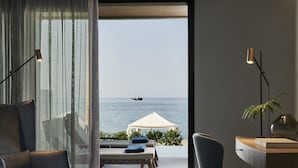 A bedroom with a large window overlooking the sea, a bed, a desk with a chair, and a small table with a plant. - AMIRA LUXURY RESORT & SPA (Rethymno)