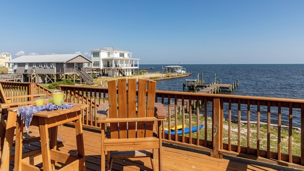 A wooden deck with a wooden chair and table, overlooking a body of water and a pier with a house in the background. - Bayfront, kayaks, private dock, 5 minute walk to the Gulf (Gulf Shores)