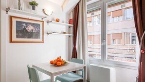 Ein Zimmer mit Esstisch, Stühlen, einem gerahmten Bild, Regalen, einem Heizkörper und einem Fenster mit Vorhängen.