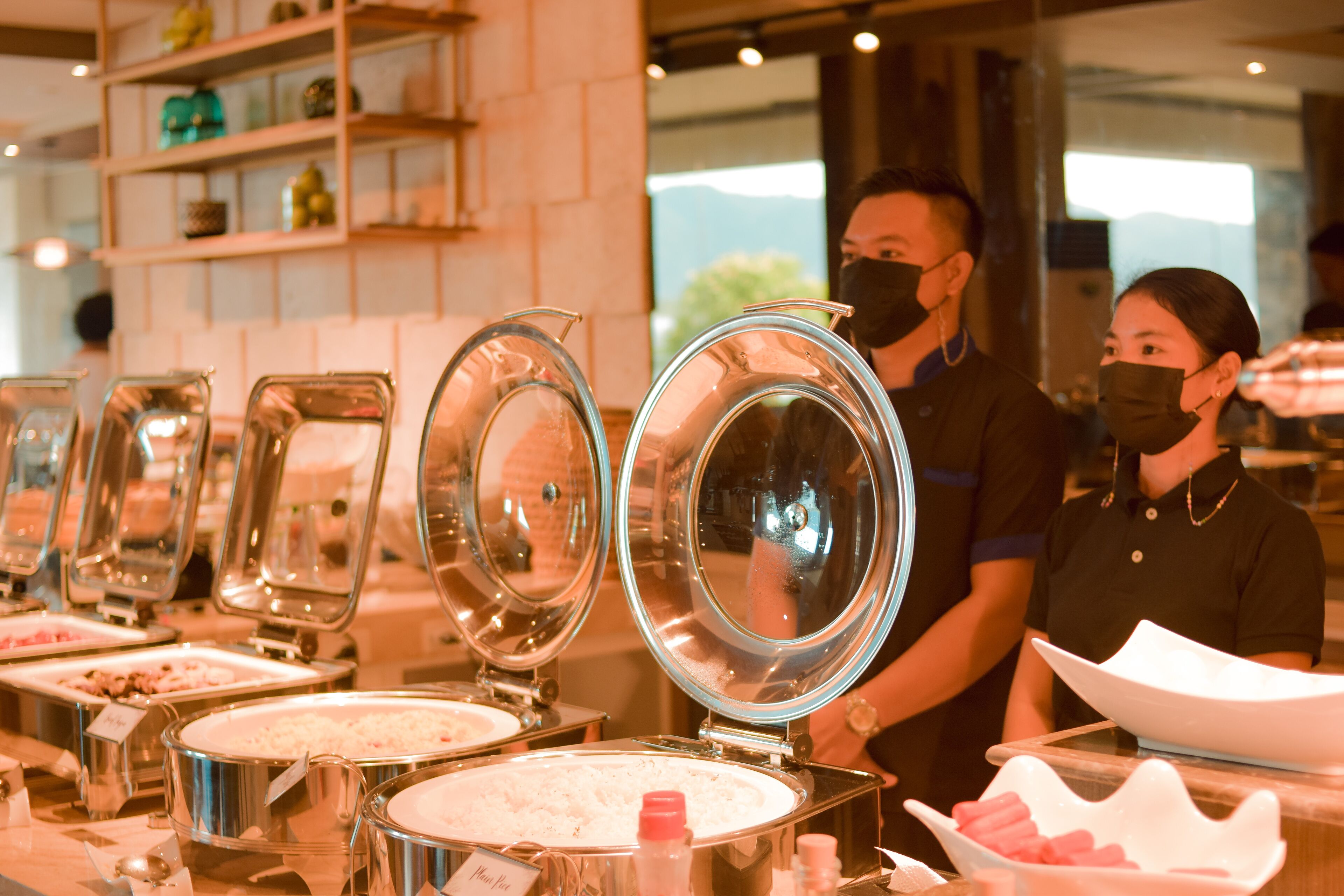 A buffet setup with multiple food dispensers, a staff member, and a customer.