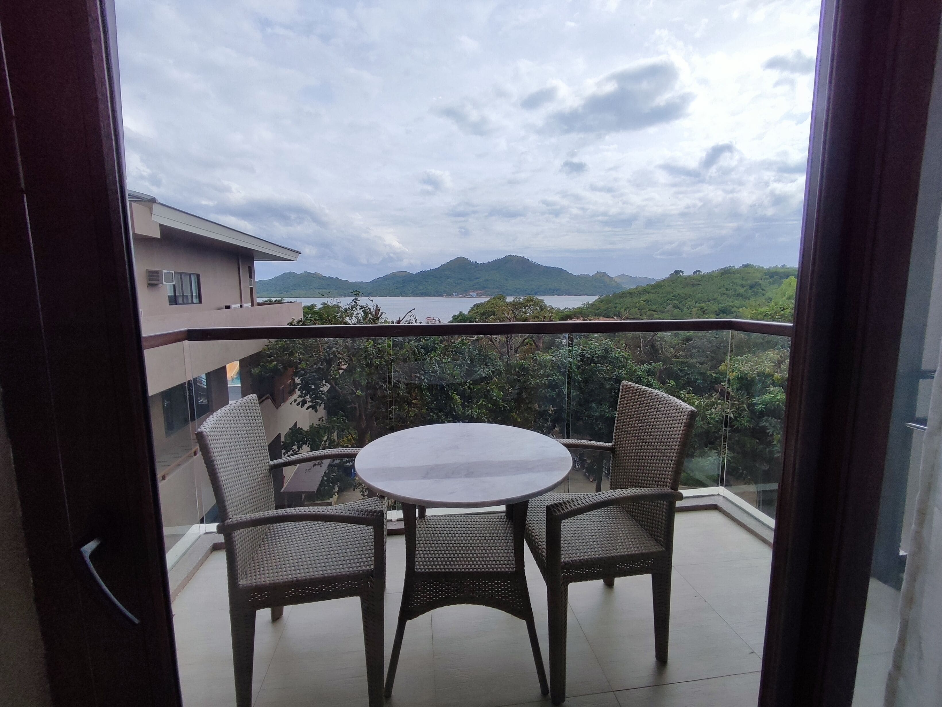 A balcony with wicker chairs and a table, overlooking a mountainous landscape and a body of water.