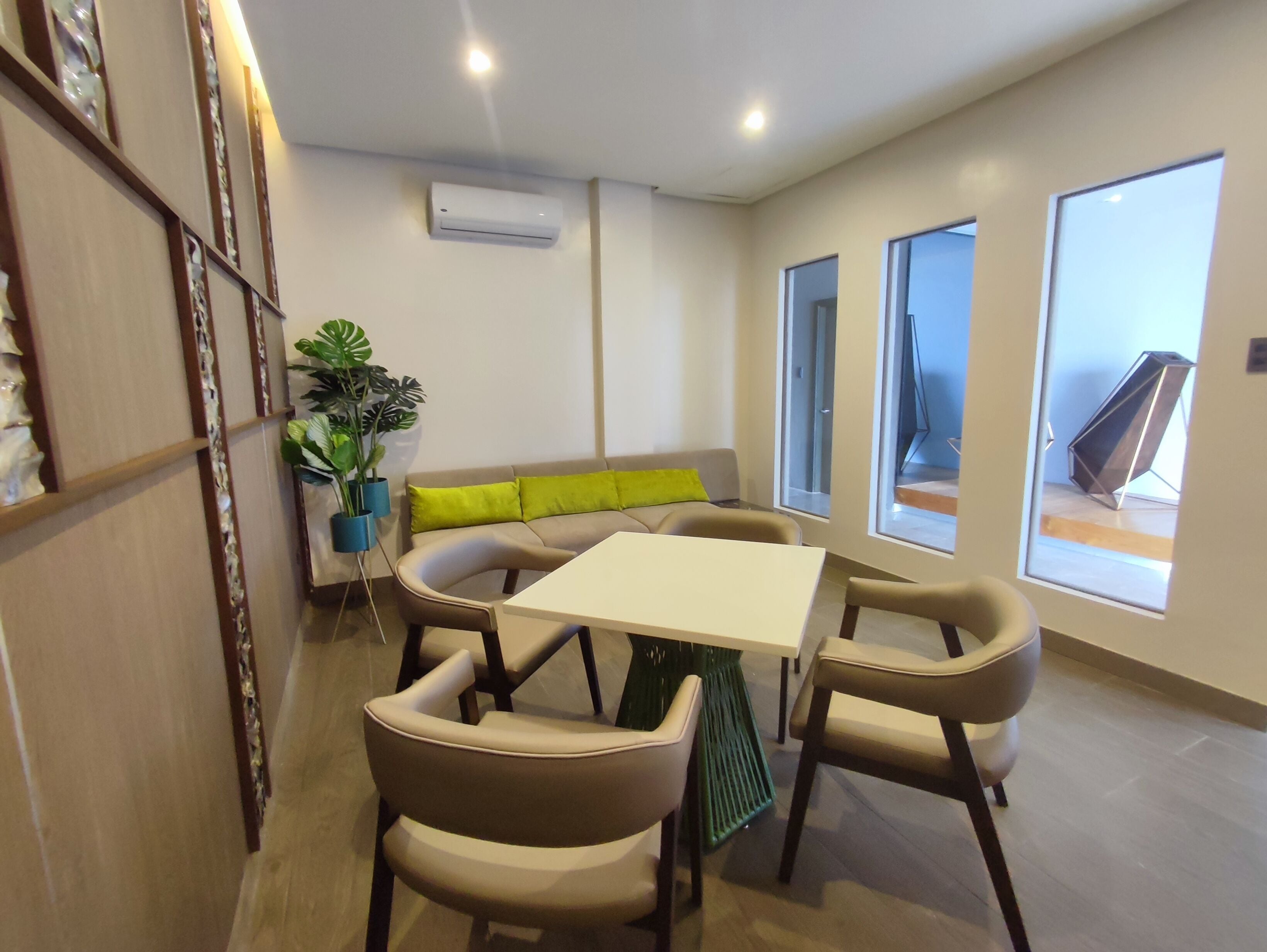 Un salon moderne avec un canapé beige, une table blanche et quatre chaises. On aperçoit une plante en pot sur la gauche et une grande fenêtre sur la droite.