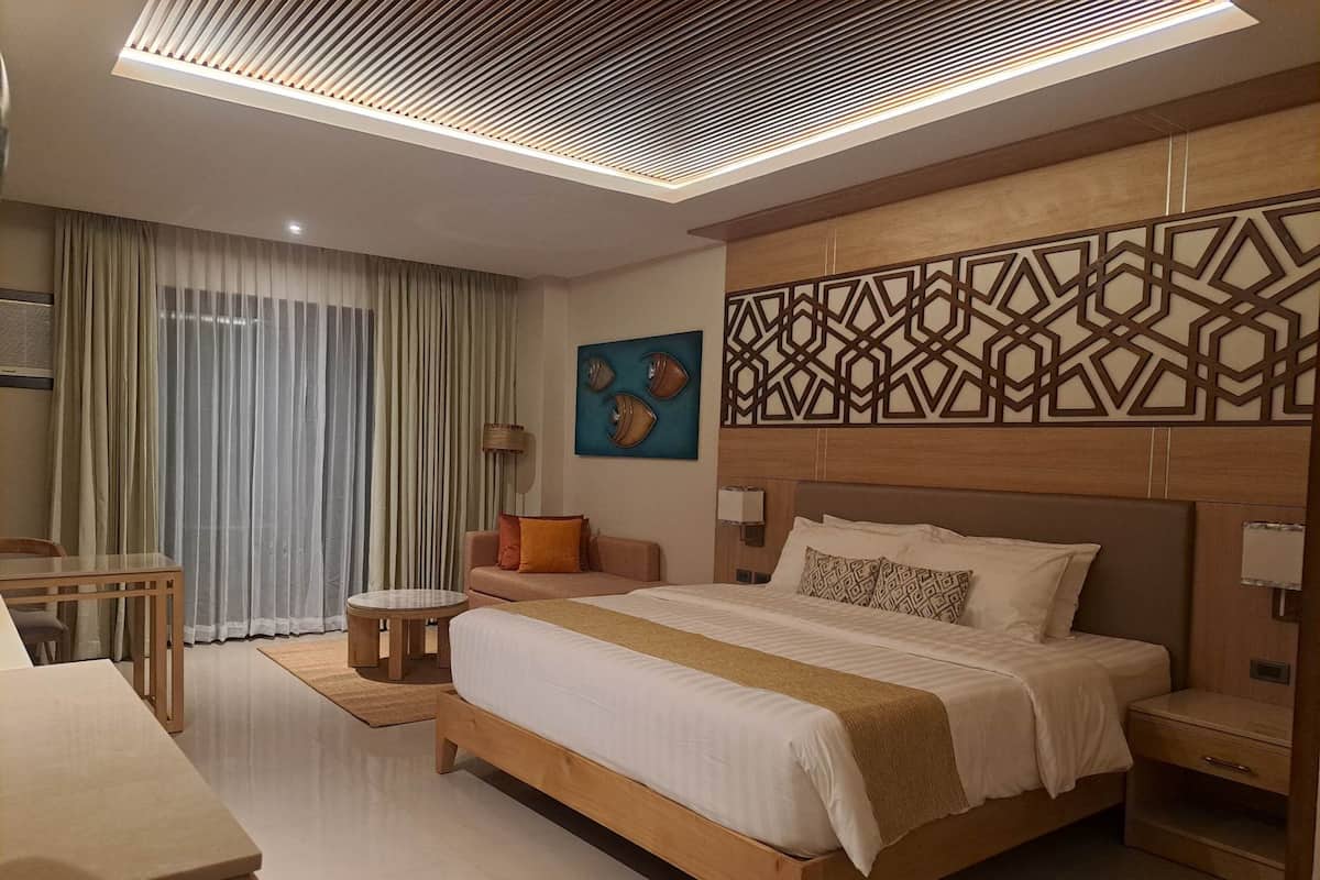 A modern hotel room with a large bed, a wooden headboard, a patterned wall decoration, a small round table, and a desk.