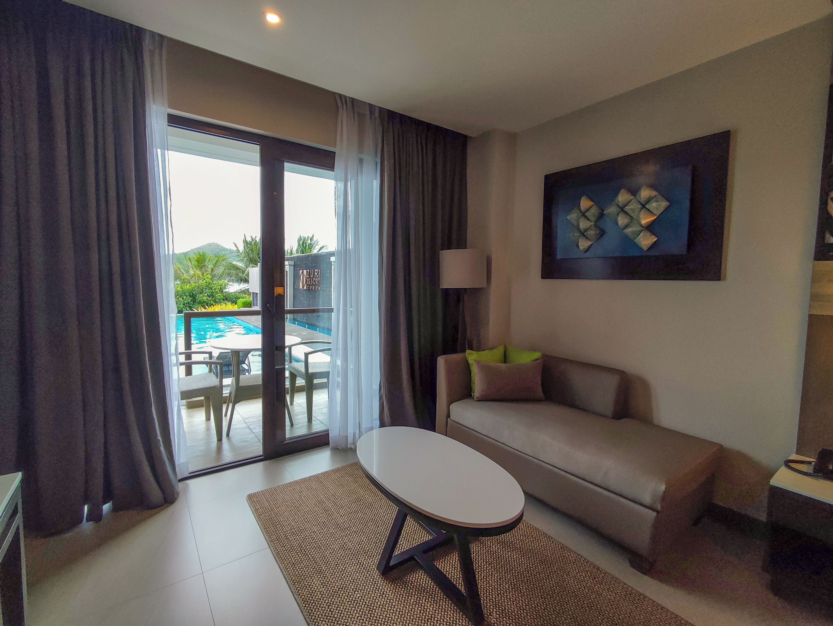 Habitación de hotel con sofá, una mesita, lámpara y vista a una piscina.