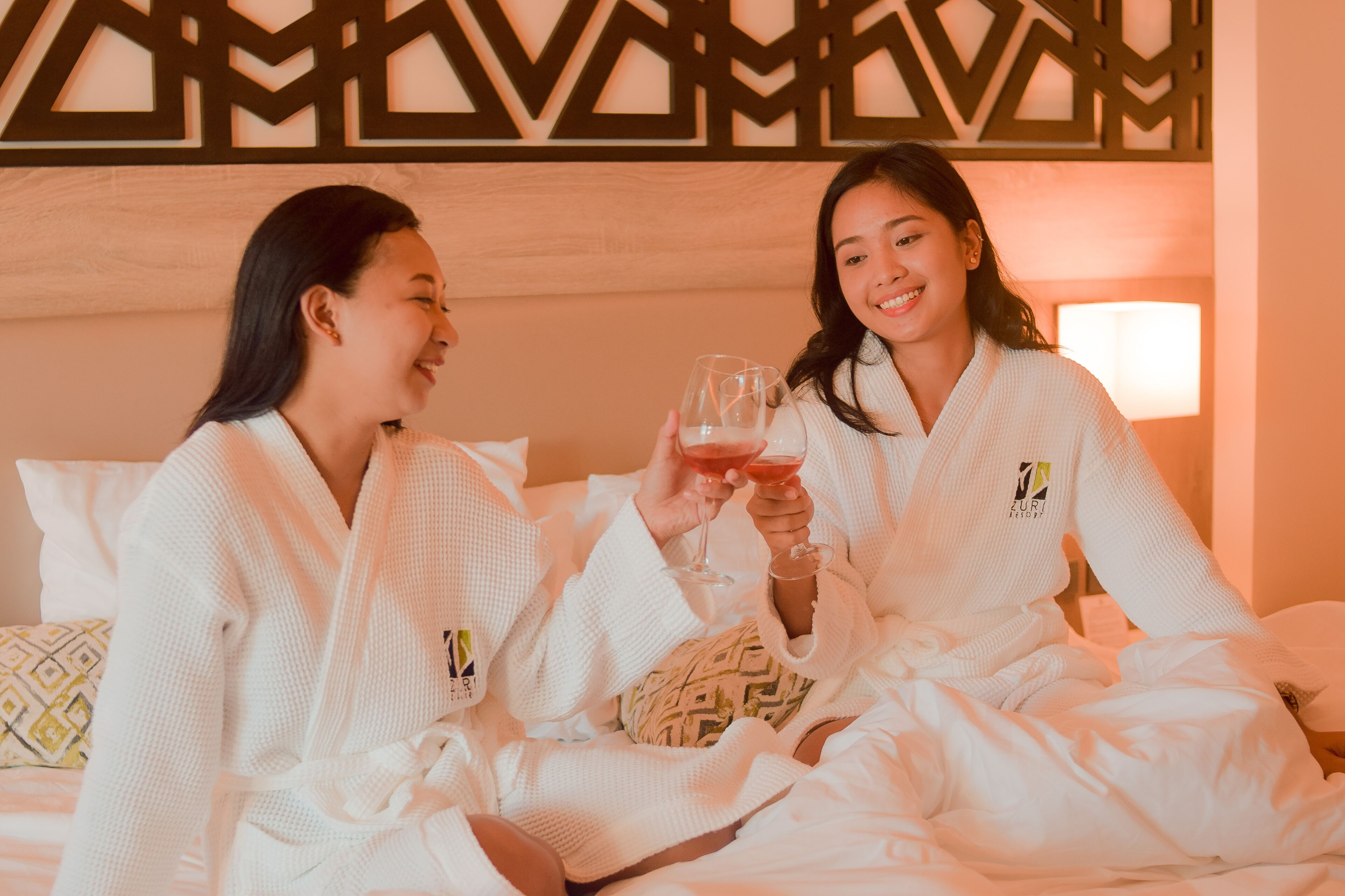 Two people in white robes sitting on a bed with a patterned pillow, holding glasses of a red drink.