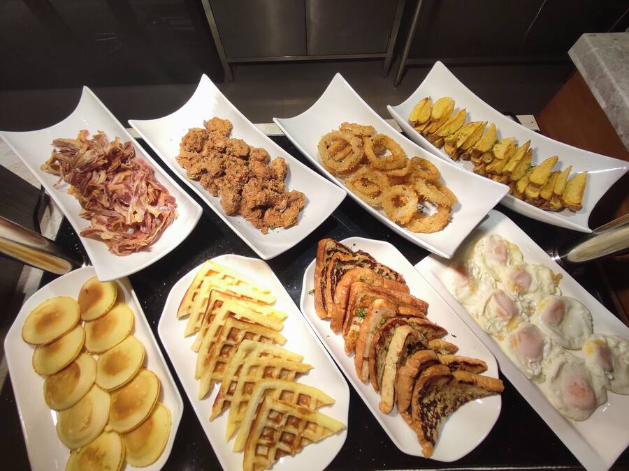 Una mesa de buffet con diversos platos, incluyendo pollo frito, aros de cebolla, alitas de papa, gofres y huevos.