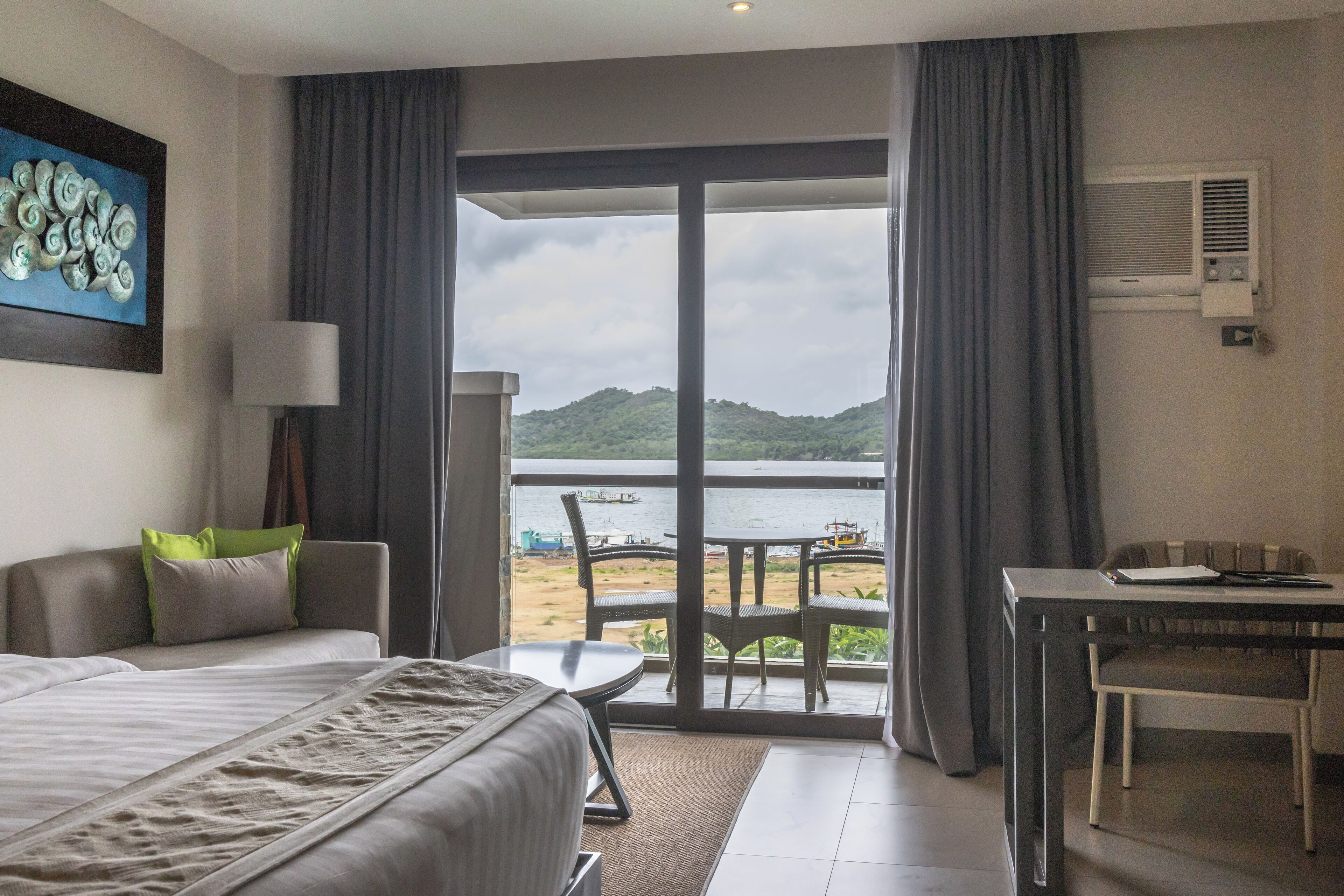 A hotel room with a bed, a desk, a chair, and a view of the beach and water.