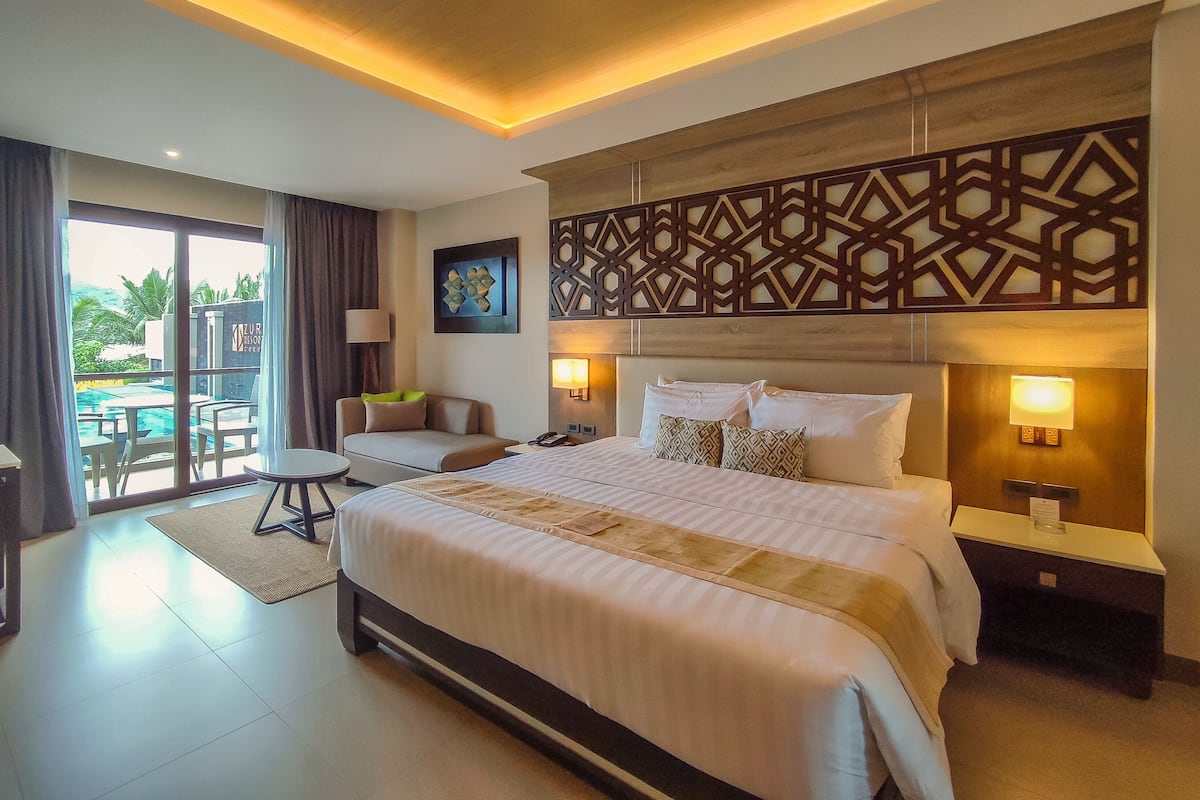 A hotel room with a large bed, a desk, a chair, a sofa, a view of the pool, and a decorative wall panel.