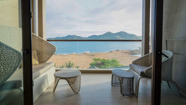 Un balcón con mobiliario de mimbre, vista a la playa y montañas al fondo.