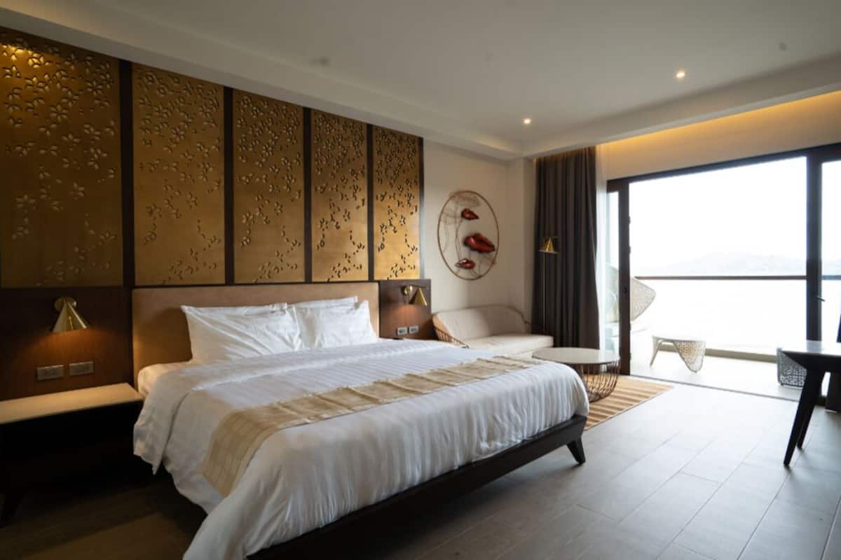 A modern hotel room with a large bed, a seating area with a sofa and armchairs, a dining table, and a large window offering a view.