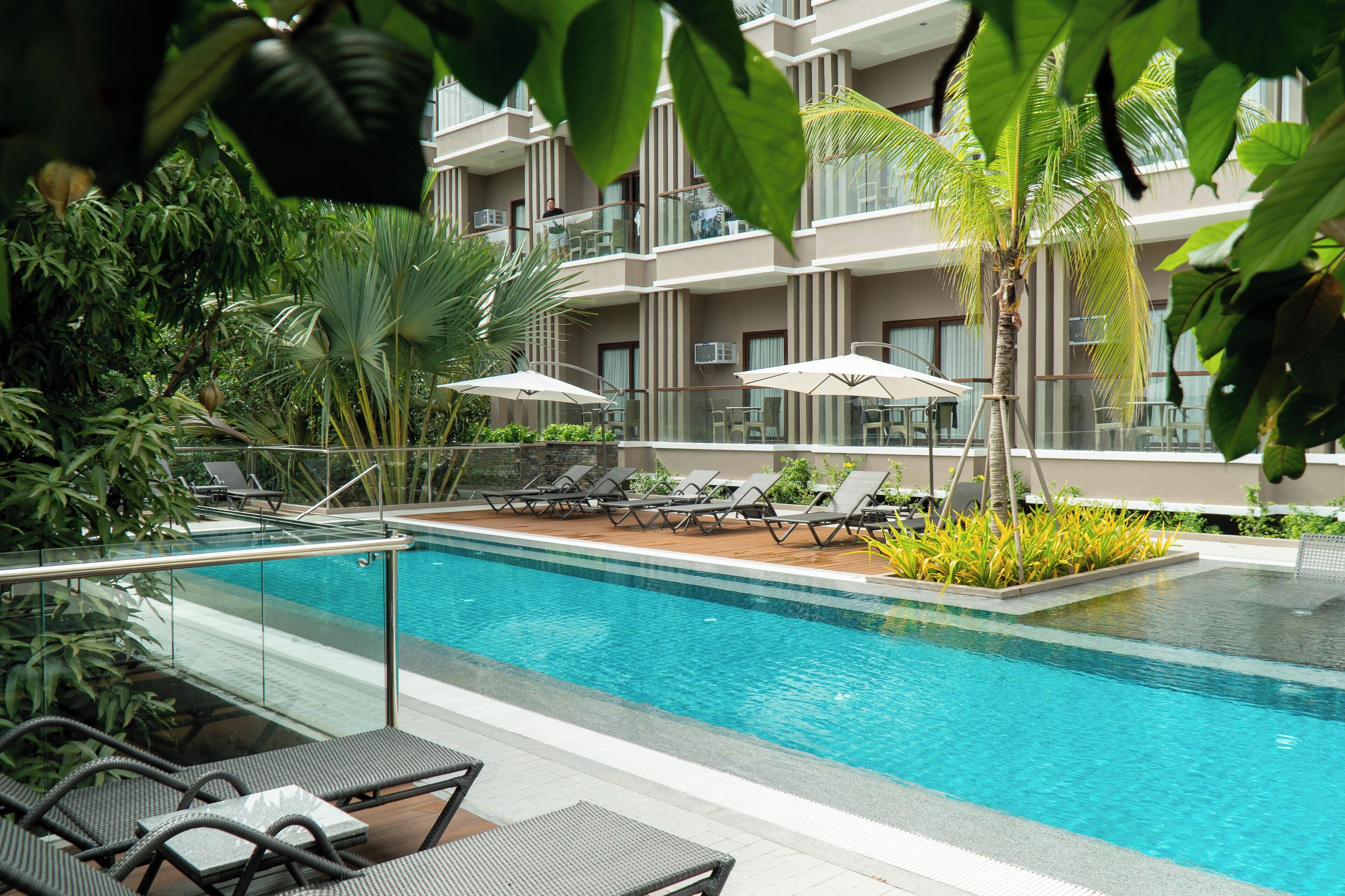 Zona de piscina con sillas de descanso y sombrillas, rodeada de vegetación exuberante y un edificio de varios pisos con balcones.