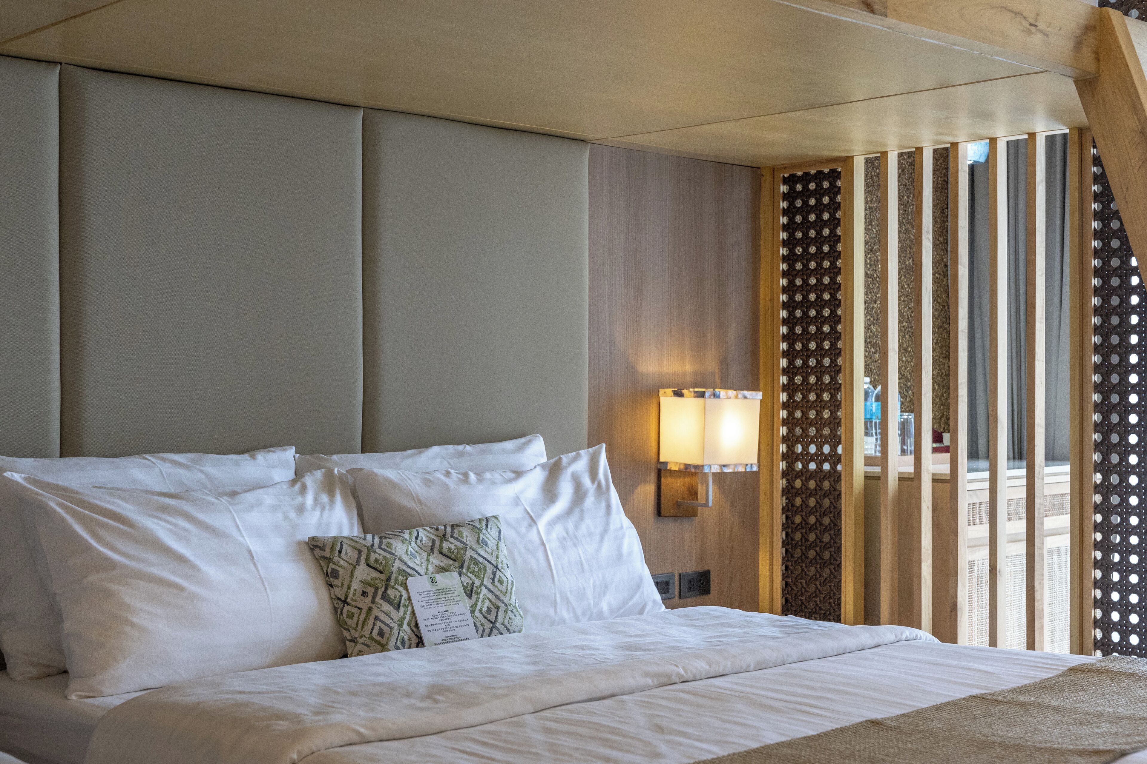 A hotel room with a large bed, a nightstand, a wall-mounted lamp, and a view through a wooden partition.