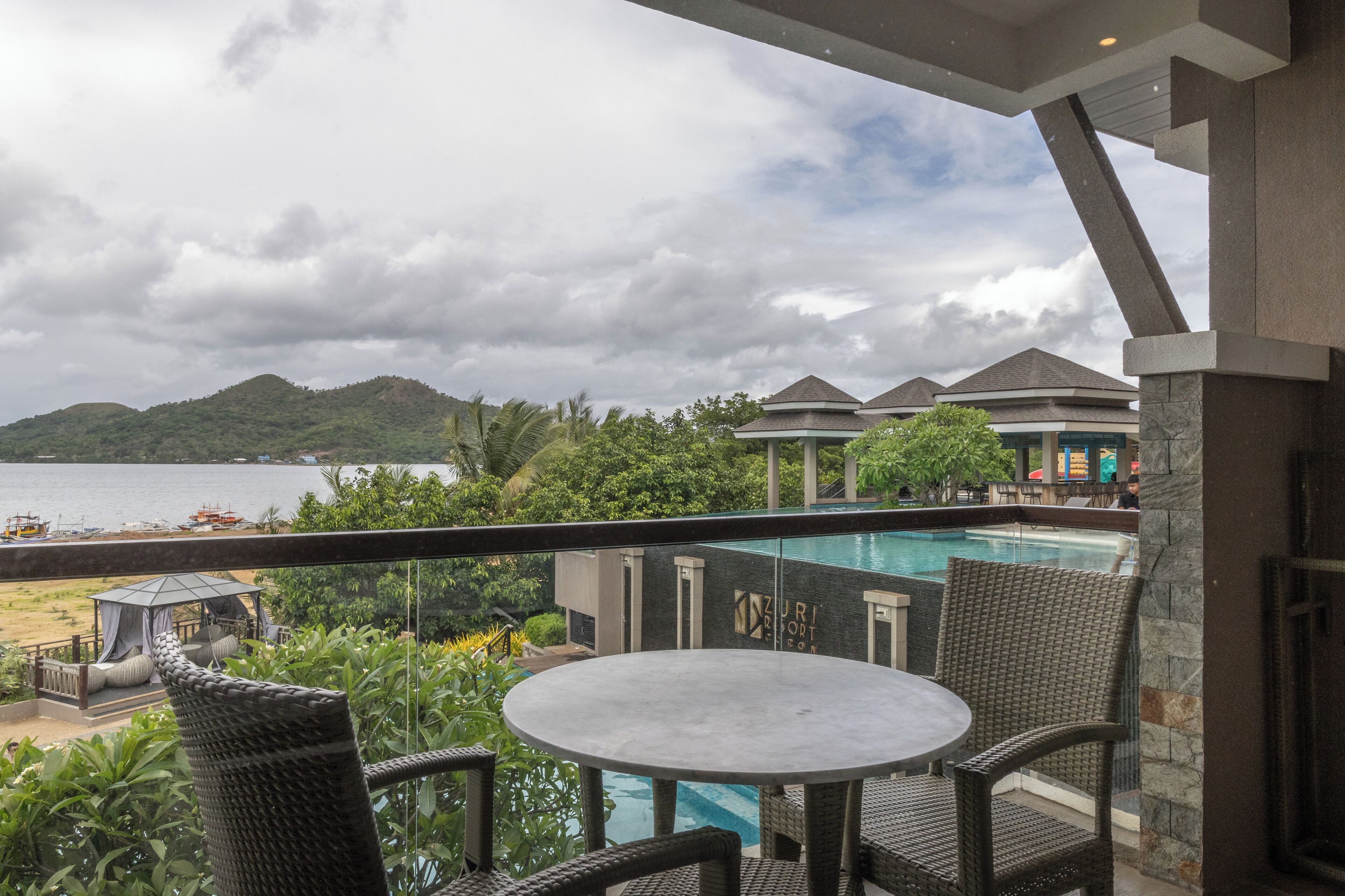 Un balcon agrémenté de fauteuils en osier et d’une table, offrant une vue sur une piscine, une plage et un paysage montagneux.