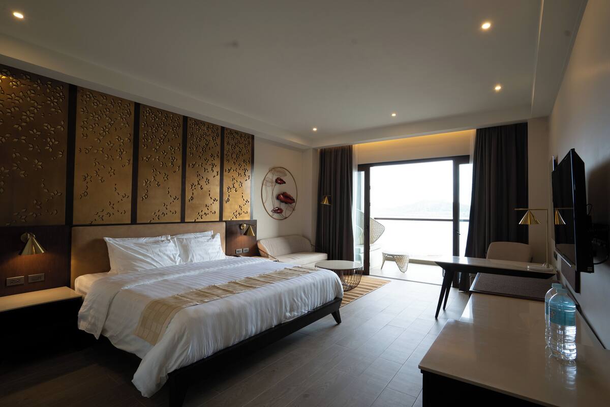 A modern hotel room with a large bed, a sofa, a desk, and a view of the outdoors.