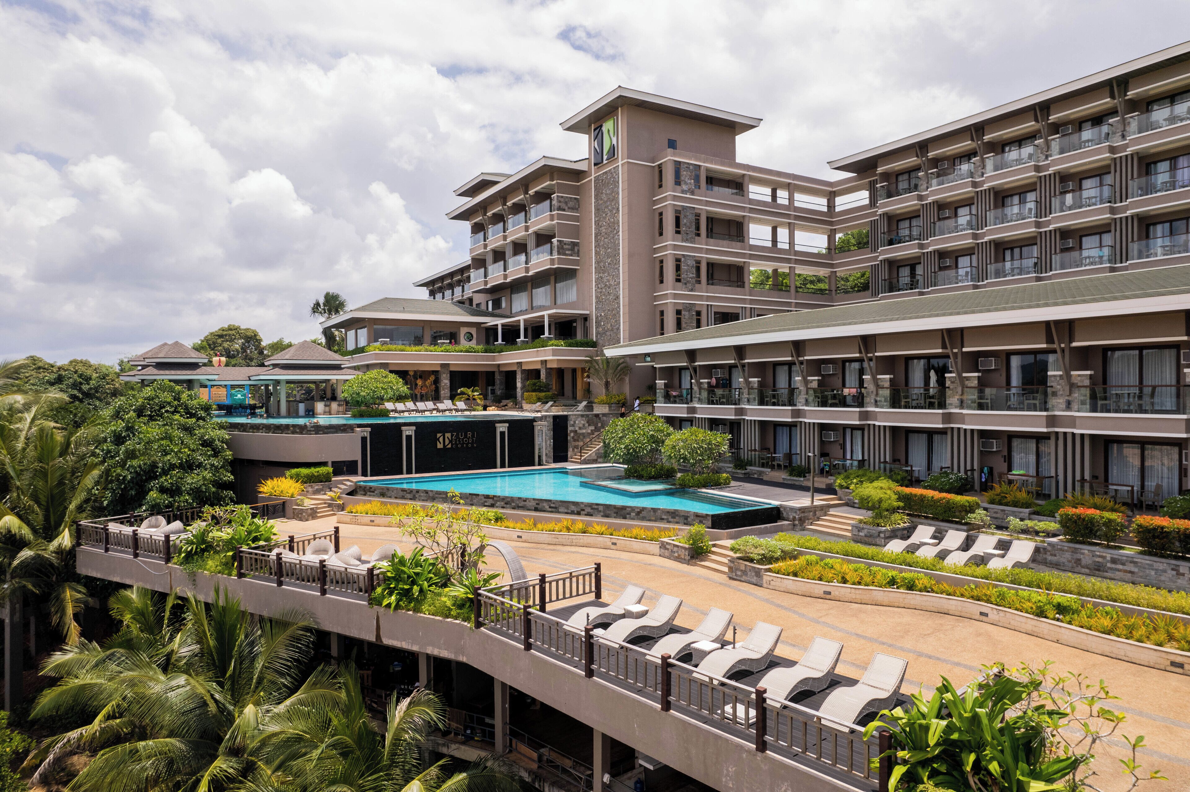 Un hotel de varios pisos con piscina, mobiliario exterior y exuberante jardinería.