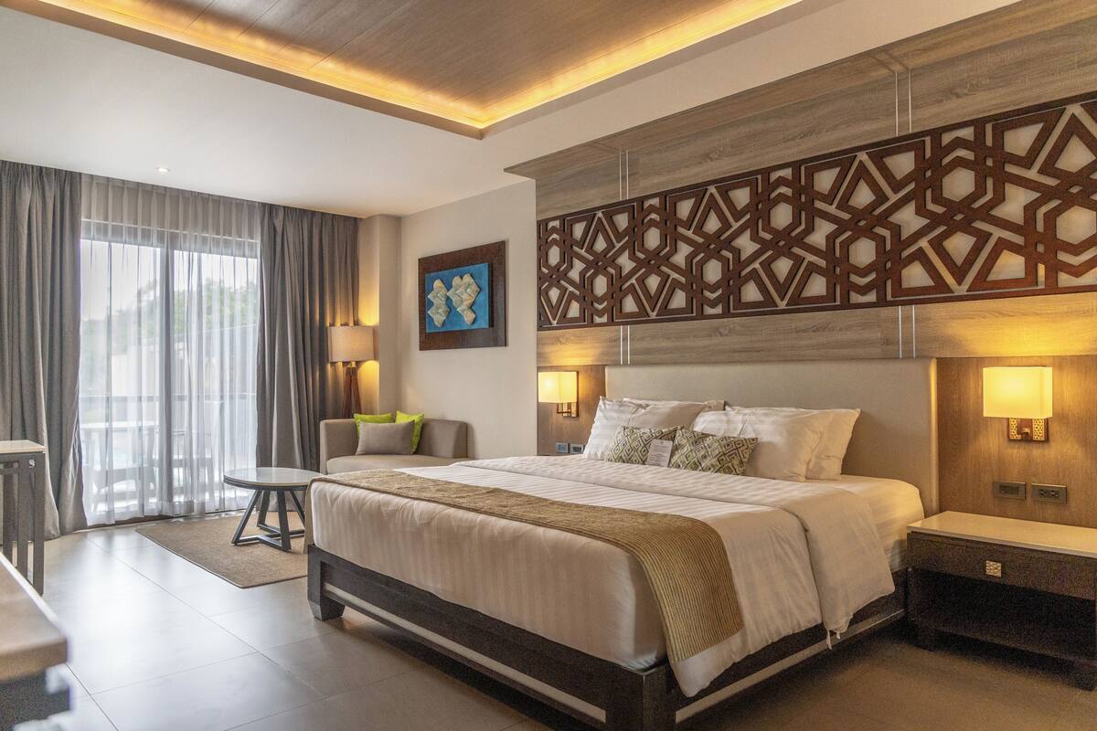 A modern hotel room with a large bed, bedside tables, a desk, and a view of the outdoors through large windows.