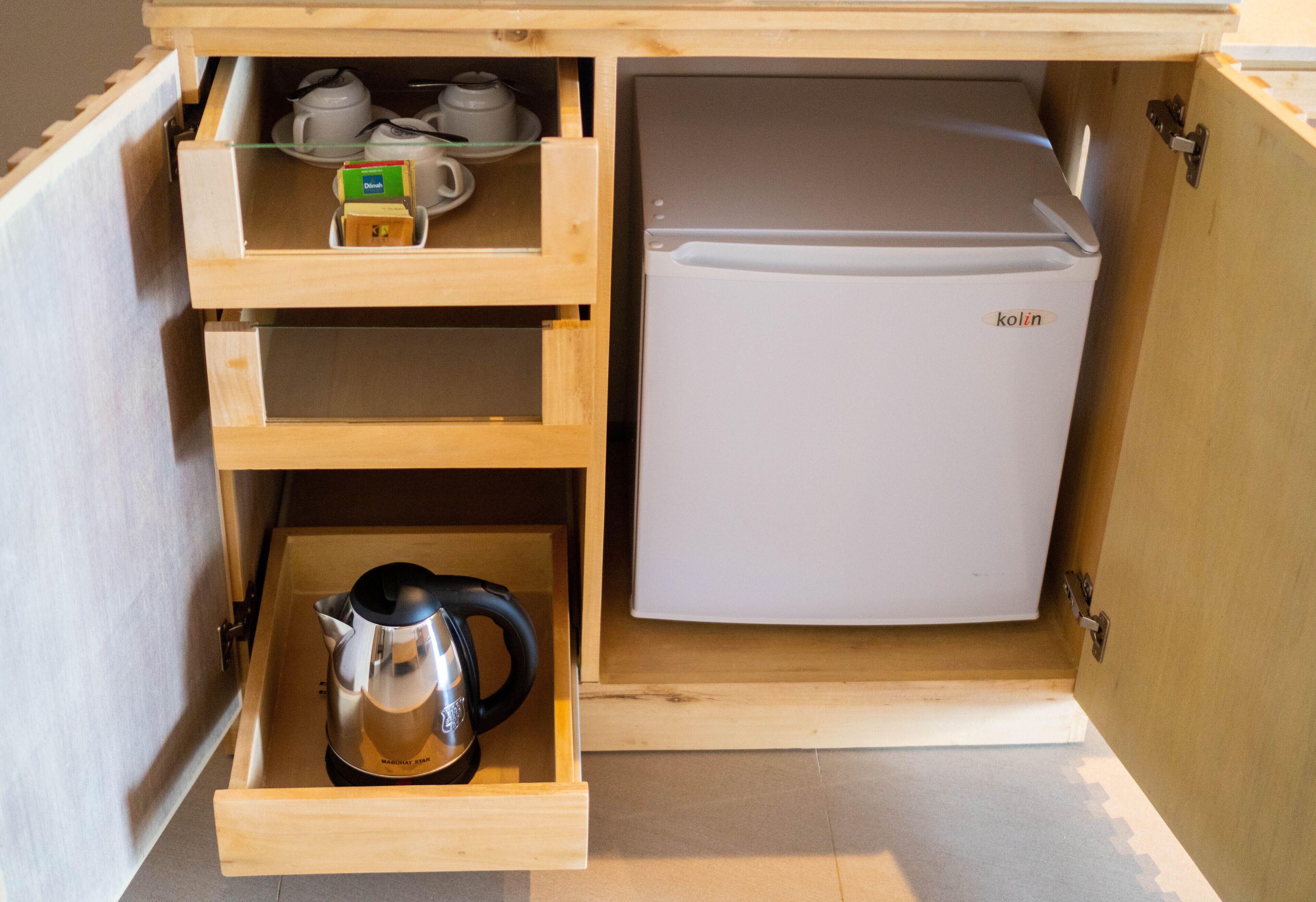 A compact kitchenette with a white refrigerator, a coffee maker, and a kettle in a wooden drawer unit.