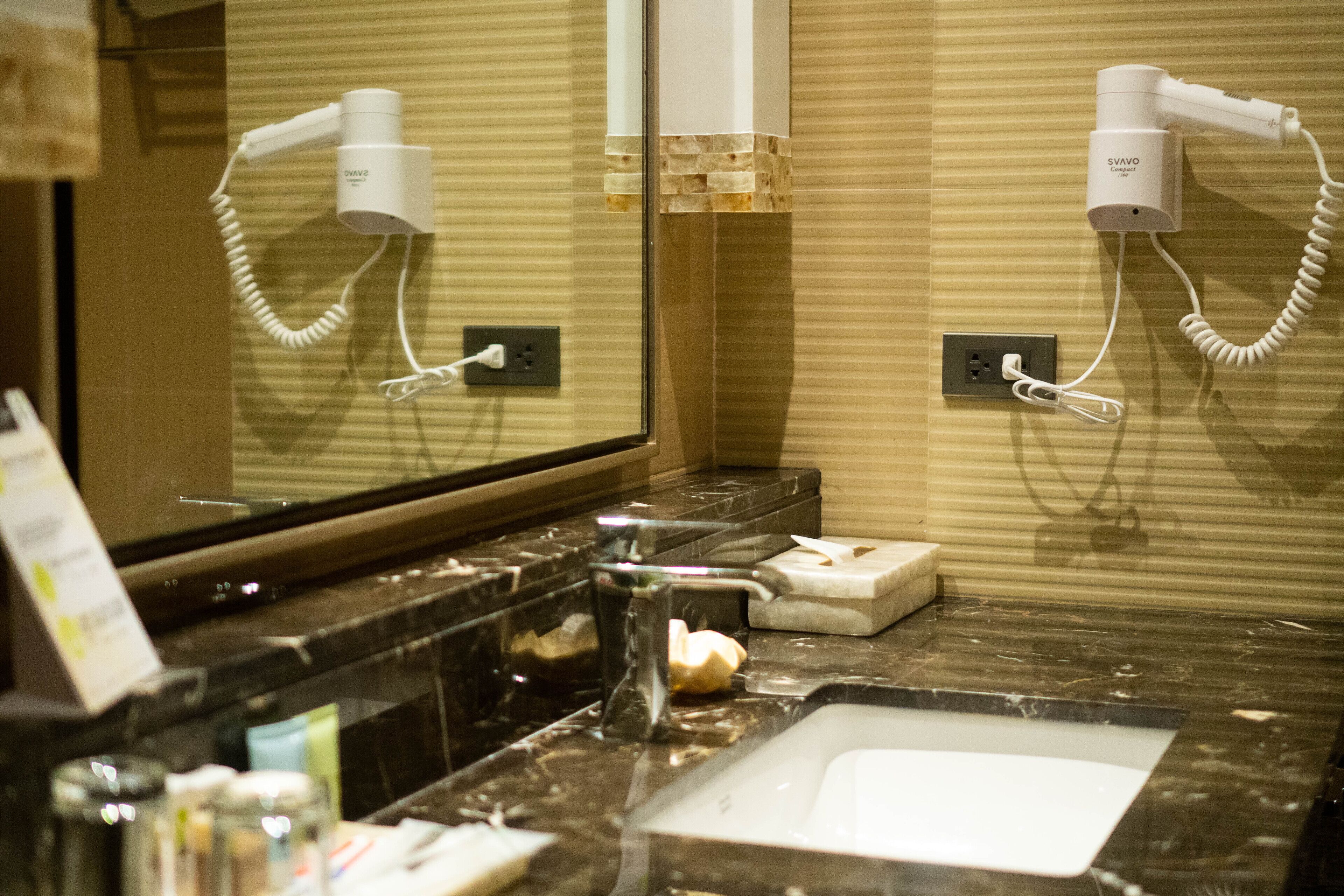 A bathroom with a large mirror, two hair dryers mounted on the wall, a sink, and a marble countertop.