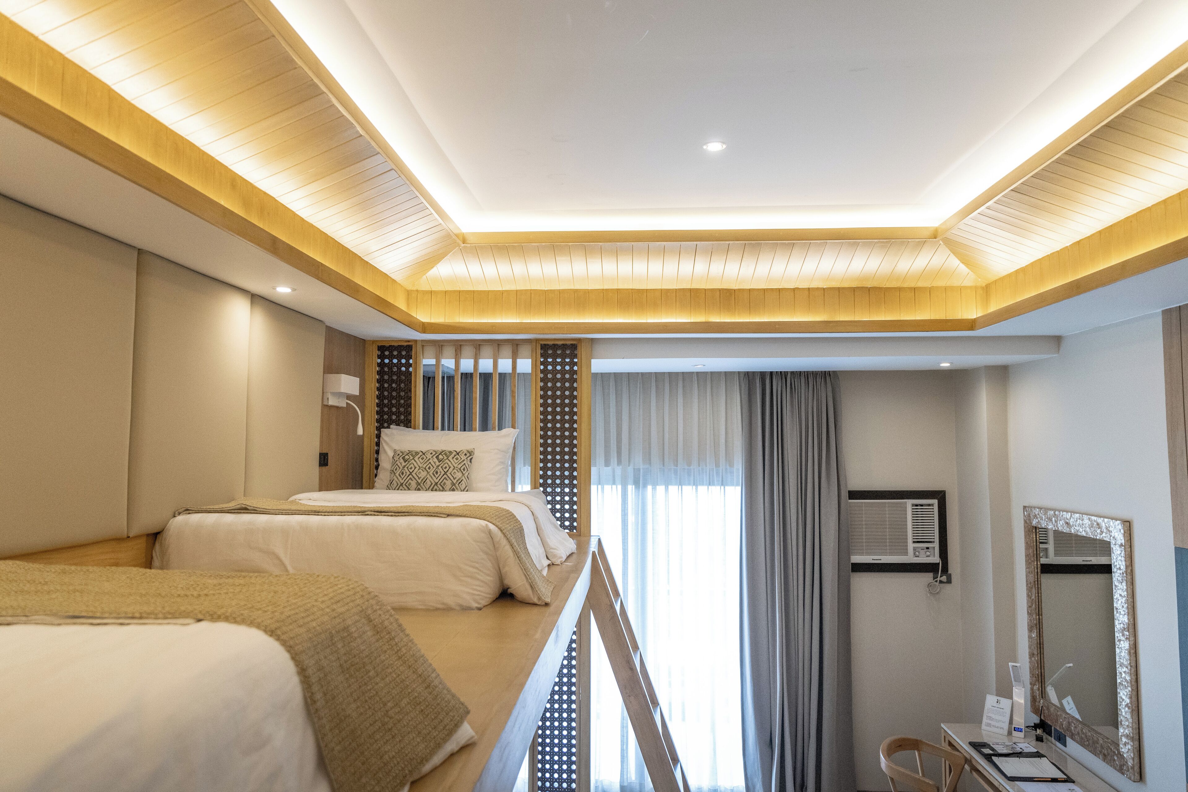 Habitación de hotel con dos camas, un escritorio con silla, un espejo grande y una ventana con cortinas.