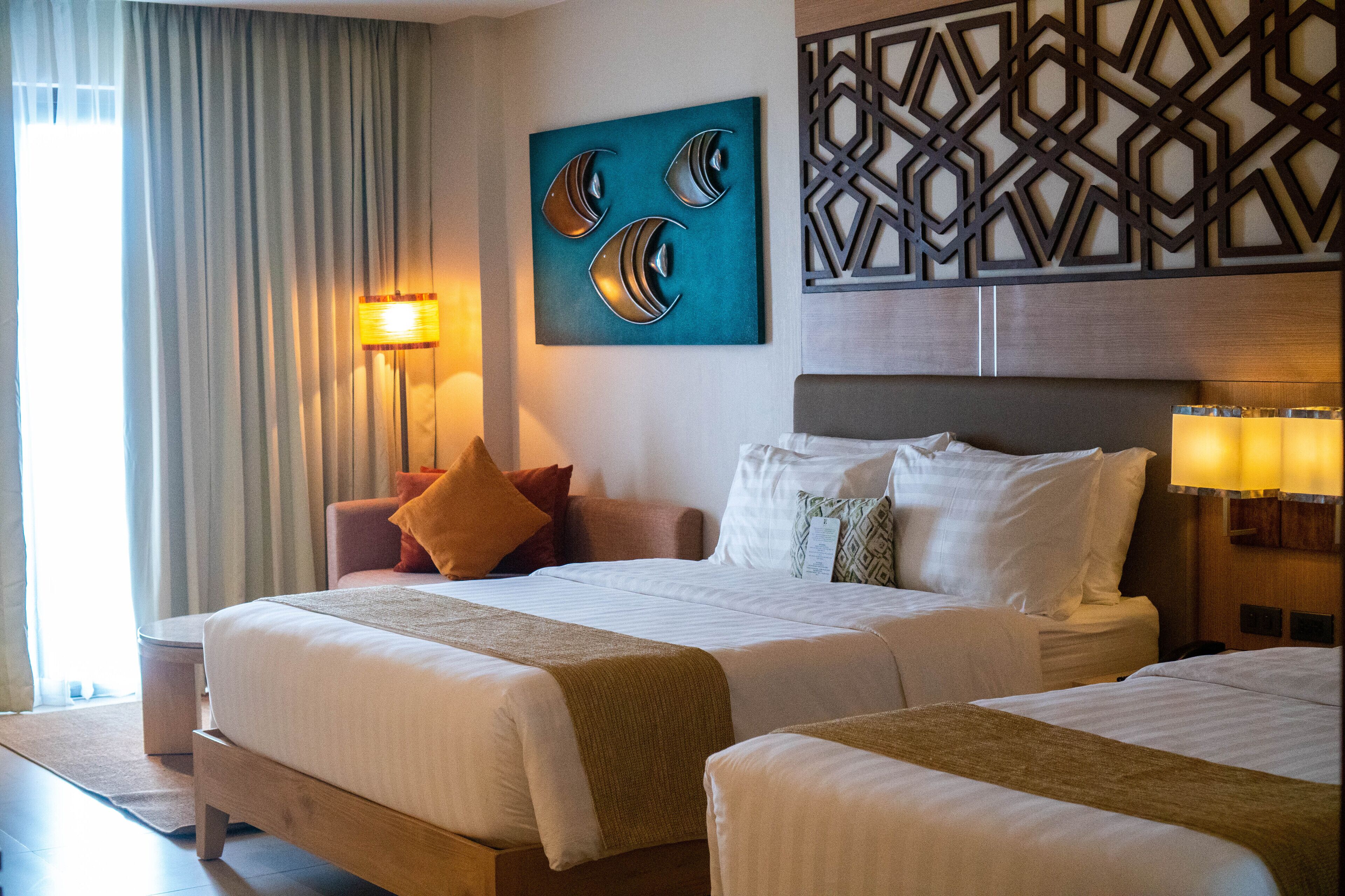 A hotel room with two beds, a large wall art piece, a geometric wall panel, and a window with curtains.