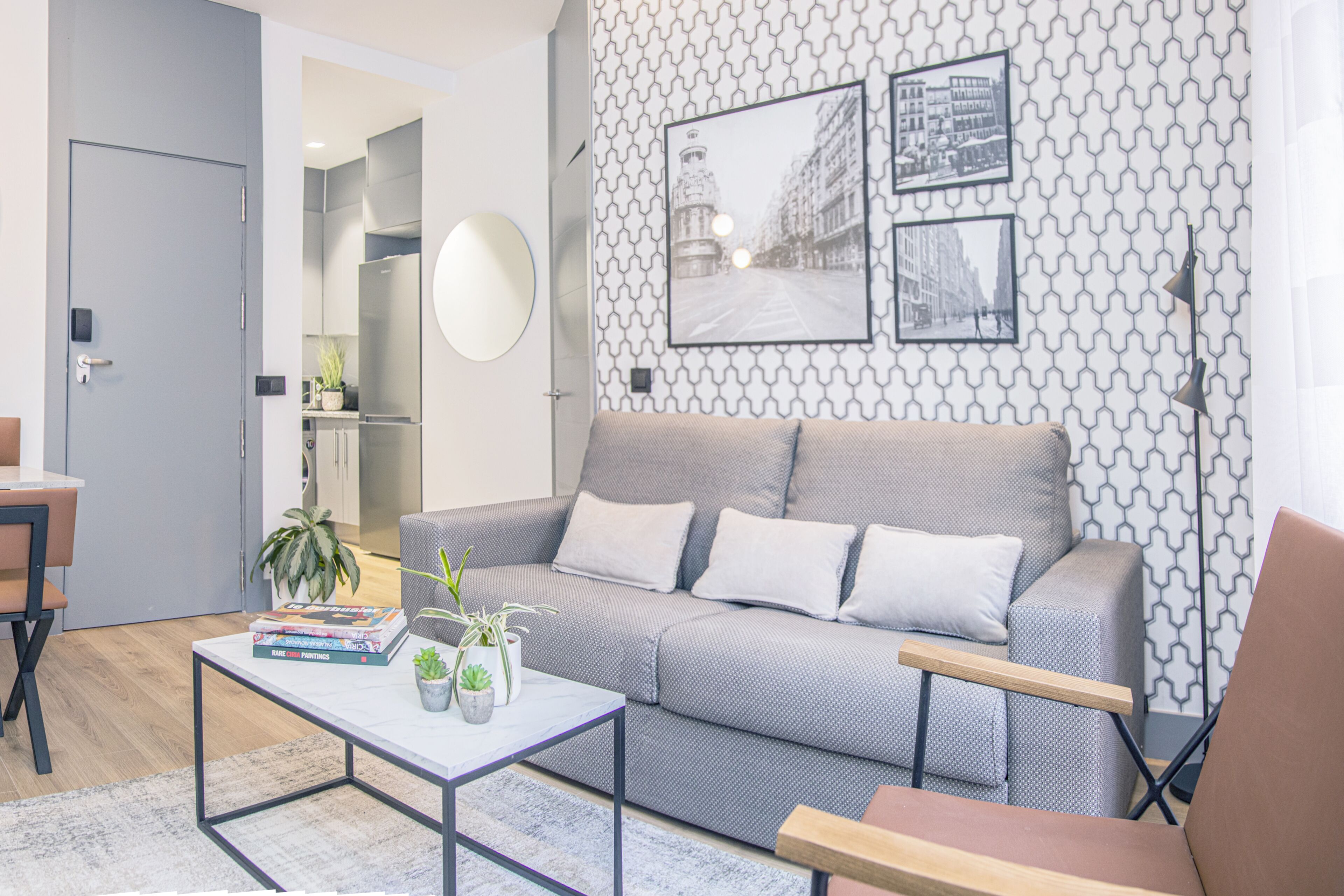 A modern living room with a grey sofa, a coffee table with books and plants, a dining area with a table and chairs, and a kitchen in the background.