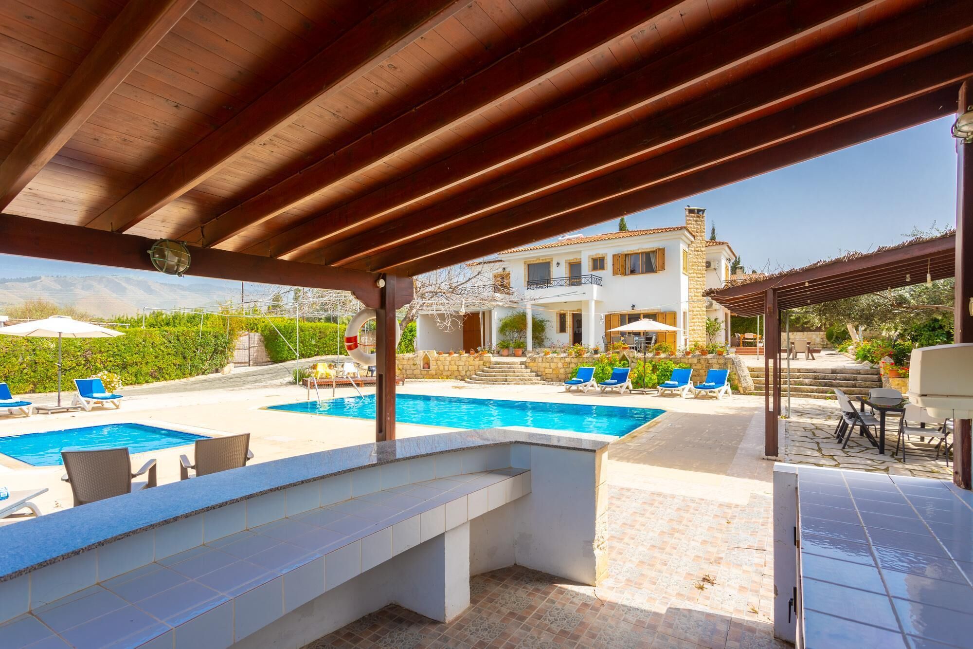 A covered outdoor swimming pool area with lounge chairs, a barbecue, and a view of a villa.