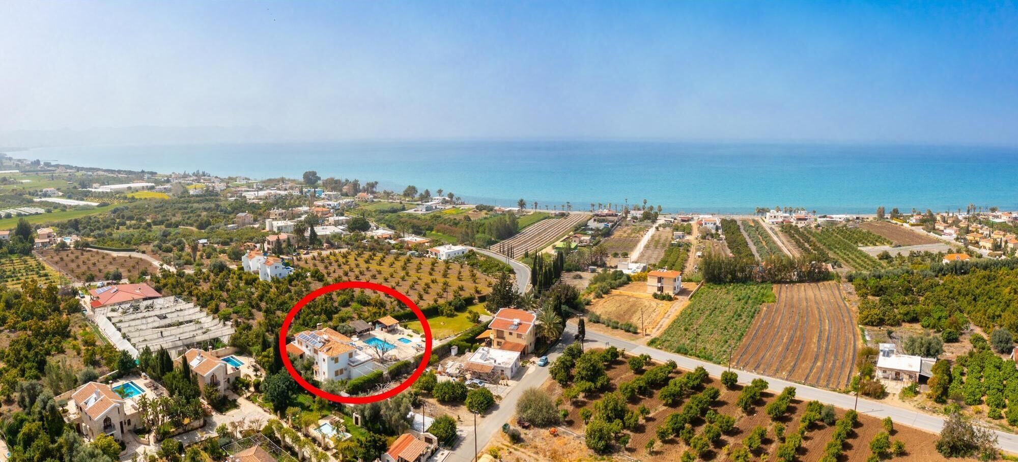 An aerial view of a coastal town with a large field of crops, a small cluster of houses with a swimming pool, and a view of the sea.