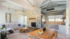 A spacious living area with a wooden dining table, comfortable seating, a stone fireplace, and a large window providing natural light. - Villa Ersi in Ionian Islands (Kefalonia)