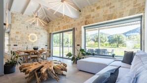 A modern living room with a stone wall, wooden furniture, and large windows offering a view of the outdoors. - Villa Ersi in Ionian Islands (Kefalonia)