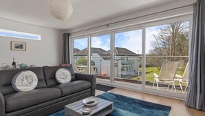 Ein modernes Wohnzimmer mit einem grauen Sofa, einem Couchtisch und einer großen Schiebeglastür, die auf einen Balkon mit Blick auf Häuser und Grünflächen führt.