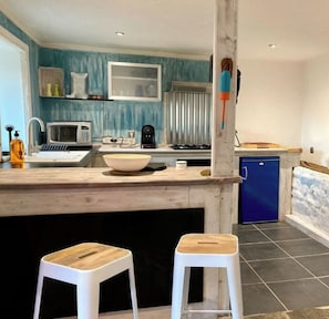 Una cocina con encimera de madera, un fregadero, un microondas, un refrigerador azul y dos taburetes blancos.