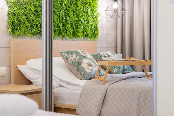 A neatly made bed with floral pillows, a bedside table with a bowl and a plate, and a green wall with a hanging plant.
