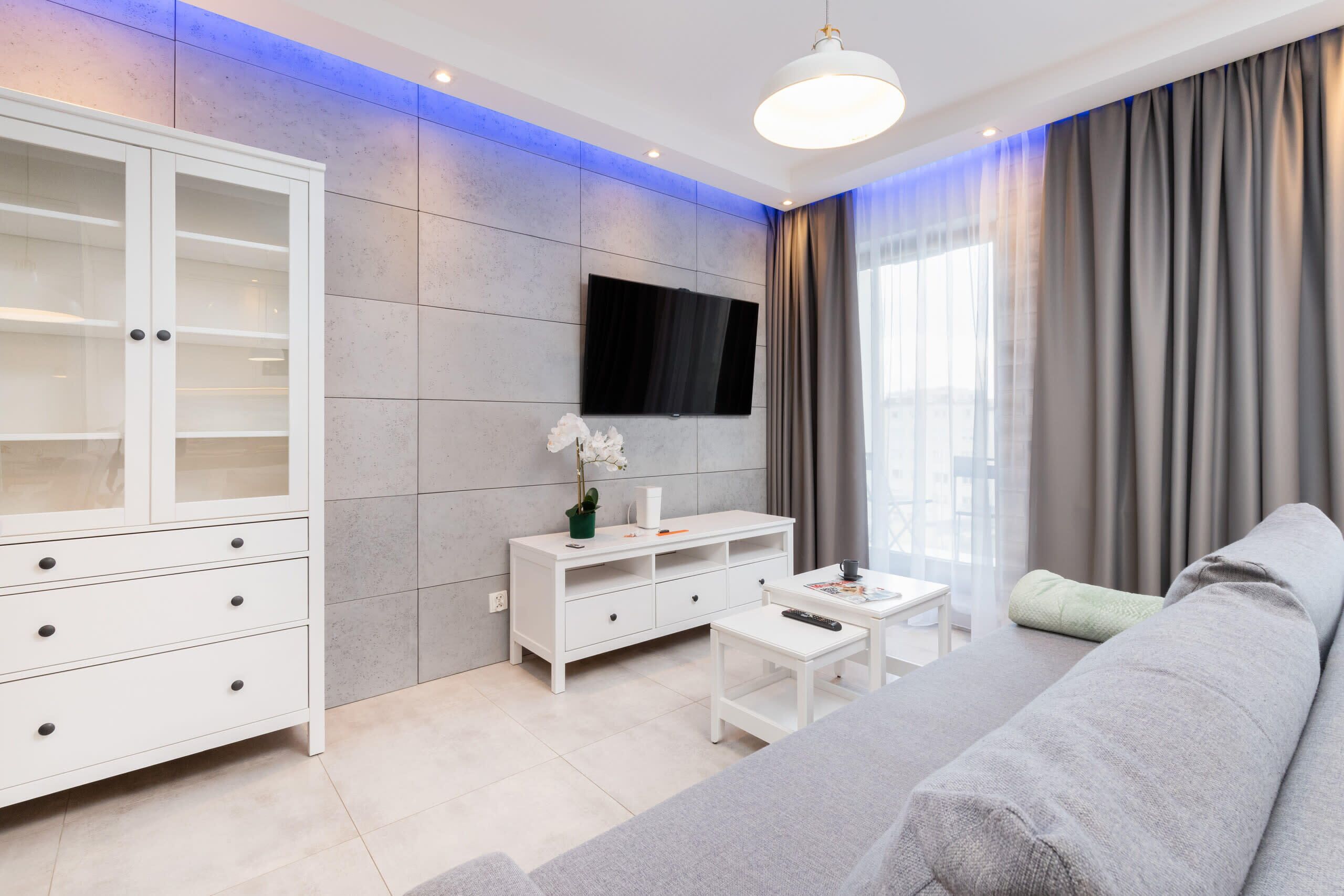 A modern living room with a grey sofa, white cabinet, flat-screen TV, and a small table with a remote control.