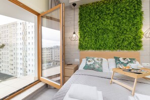 A modern bedroom with a green wall, a bed with pillows, a bedside table with a magazine, and a window overlooking a cityscape.